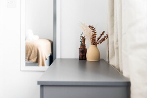 Ein Nachttisch mit einer Vase und einer Flasche, ein Spiegel, der ein Bett reflektiert, und ein Fenster mit Vorhängen.