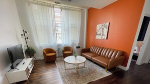 Un salon moderne avec un canapé en cuir marron, deux fauteuils orange, une table basse blanche et un téléviseur à écran plat posé sur un meuble blanc.