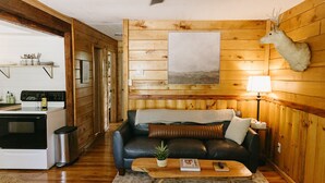 A cozy cabin interior with a wooden wall, a mounted deer head, a sofa, a coffee table, and a kitchen area with a stove and sink.