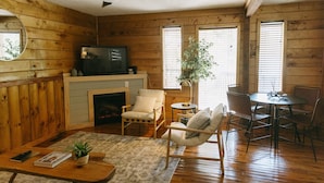 A cozy living room with wooden walls, a fireplace, a television, a dining table with chairs, and a large window with blinds.