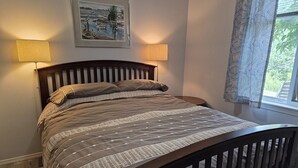 A bedroom with a wooden bed, two bedside lamps, a framed picture on the wall, and a window with curtains. - Charming walkout basement patio suite - ocean views. (Cowichan Bay)