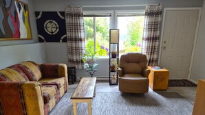 A living room with a sofa, armchair, and coffee table. There is a window with curtains, a lamp, and a door. - Charming walkout basement patio suite - ocean views. (Cowichan Bay)