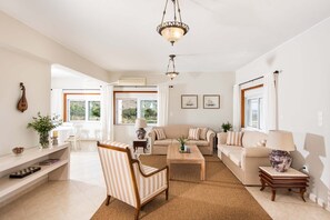 A spacious living room with a sofa, armchair, and coffee table, a chandelier, and large windows with curtains.