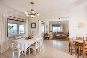 A spacious living area with a dining table, chairs, a sofa, and a coffee table. There are large windows offering a view of the outdoors.