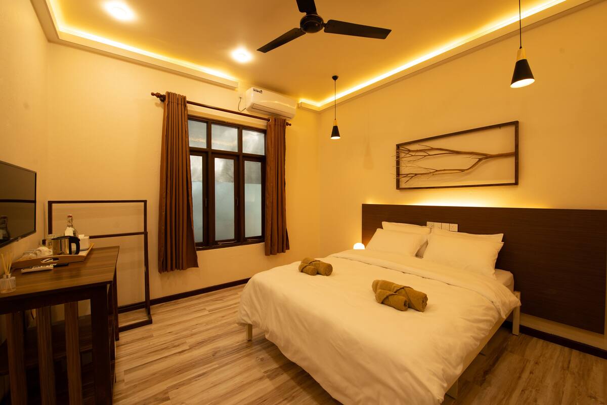 A hotel room with a bed, a ceiling fan, a window with curtains, a wall-mounted air conditioner, a bedside table with a bottle and a tray, and a painting on the wall.