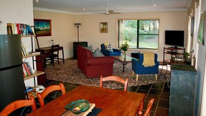 Un salón con un sofá rojo, sillones azules, una mesa de centro de madera, una estantería, un televisor y un ventanal con vistas a árboles.