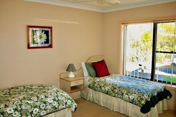 A bedroom with two single beds, a nightstand with a lamp, a framed picture on the wall, and a large window with a view of greenery. - Serena Cottages Beechworth - Your Country Getaway - 1 (Beechworth)