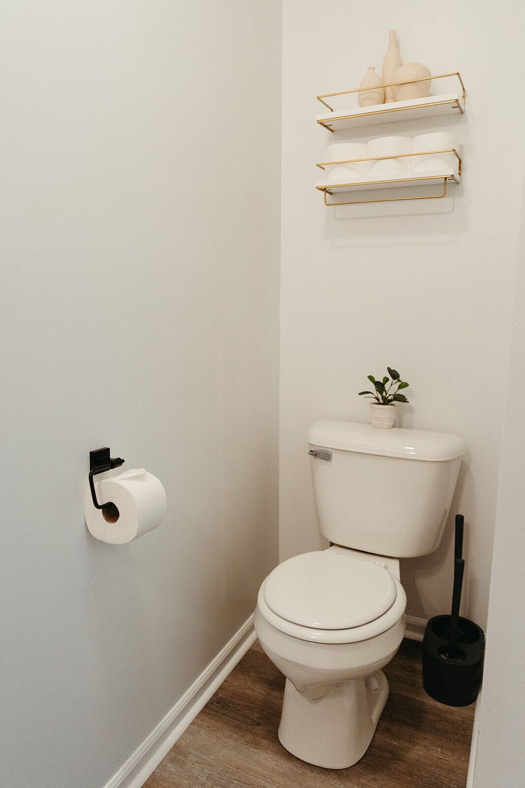 Un baño pequeño y blanco con inodoro, dispensador de papel higiénico, una planta pequeña sobre el tanque del inodoro y un cubo de basura negro.