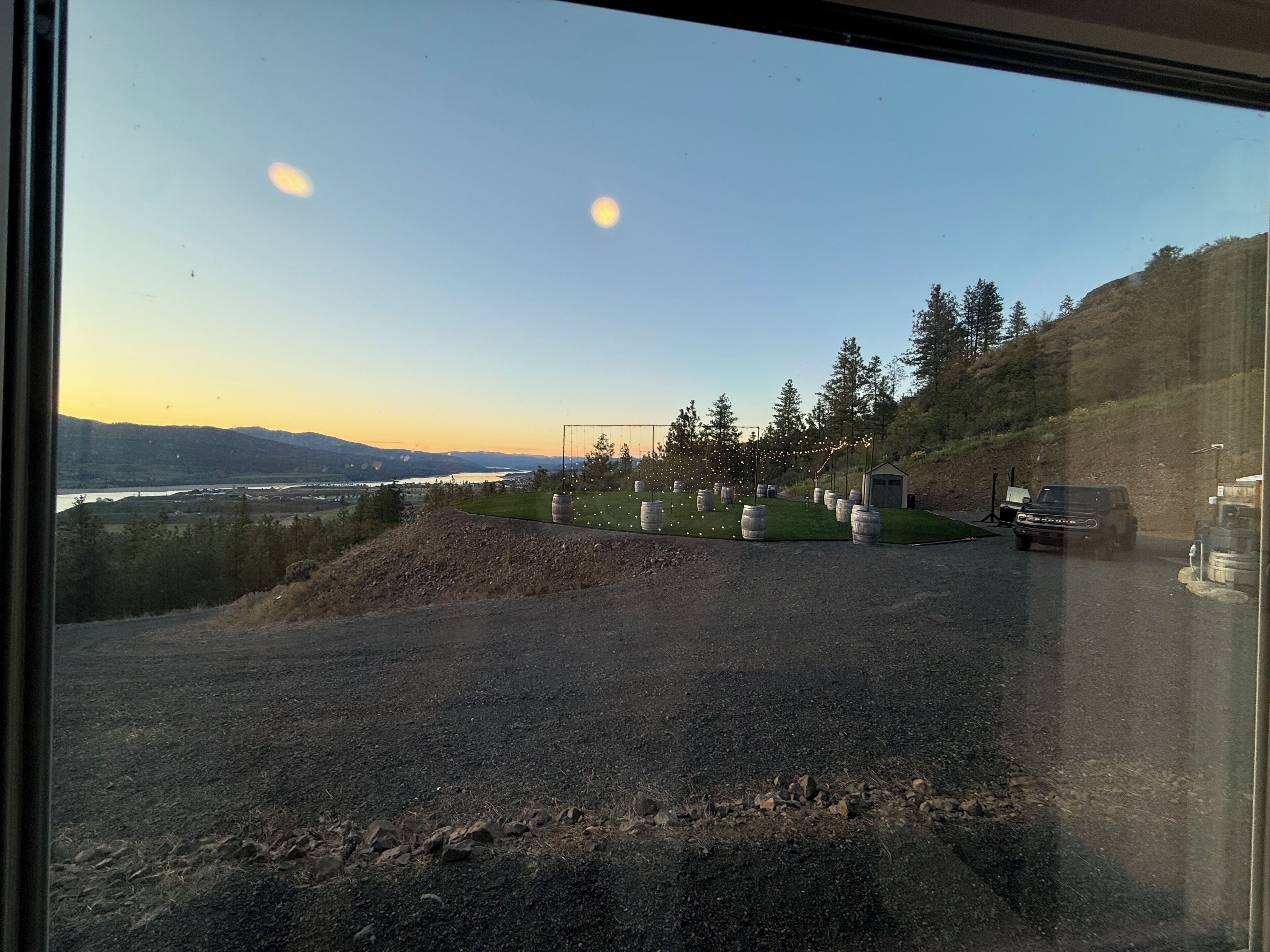 A gravel driveway leading to a building with a small shed, surrounded by trees and a view of a river and mountains.