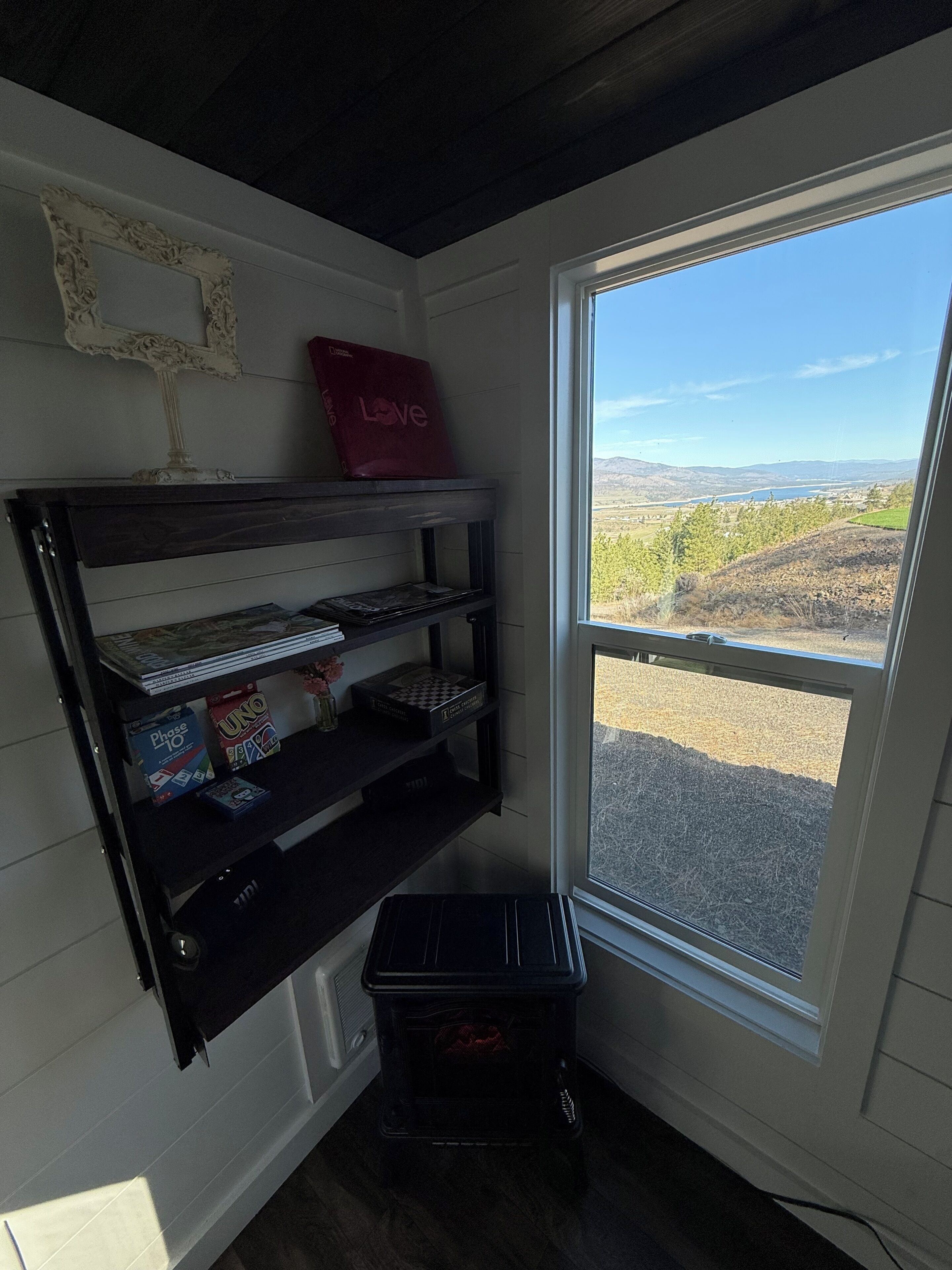 Ein Zimmer mit Ecke, ein Fenster, eine Holzablage mit Gesellschaftsspielen, ein kleiner schwarzer Heizlüfter und ein dekoratives Element an der Wand.