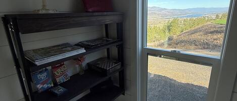 Ein Zimmer mit Ecke, ein Fenster, eine Holzablage mit Gesellschaftsspielen, ein kleiner schwarzer Heizlüfter und ein dekoratives Element an der Wand.
