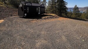 A small, black cabin with a metal roof and a door, surrounded by a gravel driveway and dense forest. - Tiny Home, Lake View, RV & Boat Parking, Pistol Range. (davenport)