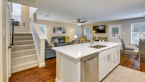 A modern kitchen with a central island, a dining area with a table and chairs, a living room with a sofa and a TV, and a staircase leading to an upper level. - Modern Woodlands Townhome w/ Spacious Yard & Patio (The Woodlands)