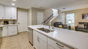 A modern kitchen with white cabinetry, a large island with a sink, and a staircase leading to a living area with a black leather sofa. - Modern Woodlands Townhome w/ Spacious Yard & Patio (The Woodlands)