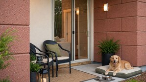 Un chien se repose sur un coussin pour animaux placé à l’extérieur d’un bâtiment doté d’une porte coulissante en verre, près d’une chaise, d’une table avec une tasse et d’une plante en pot.
