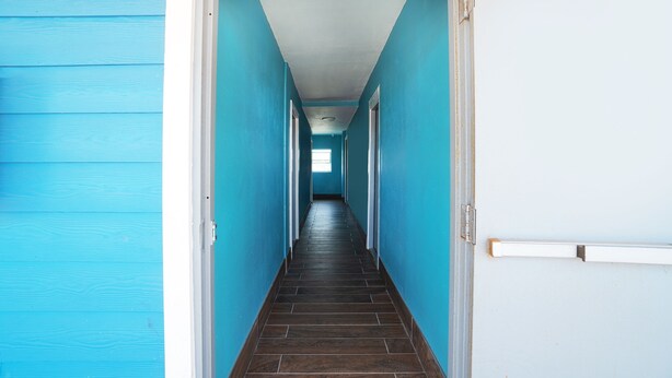 A hallway with blue walls and a dark wooden floor, leading to a door with a light switch on the wall. - Plaza Motel Corpus Christi By Oyo (Corpus Christi)