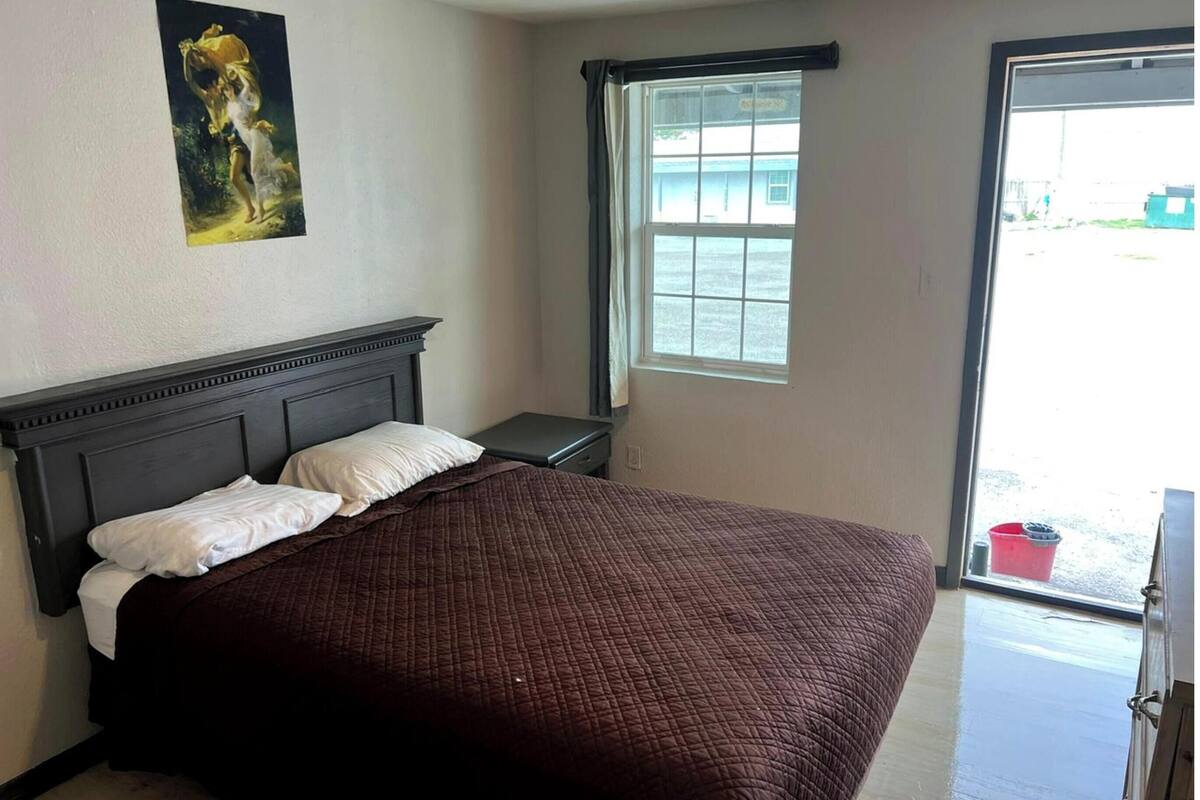 A bedroom with a bed, a nightstand, a window with curtains, and a painting on the wall.