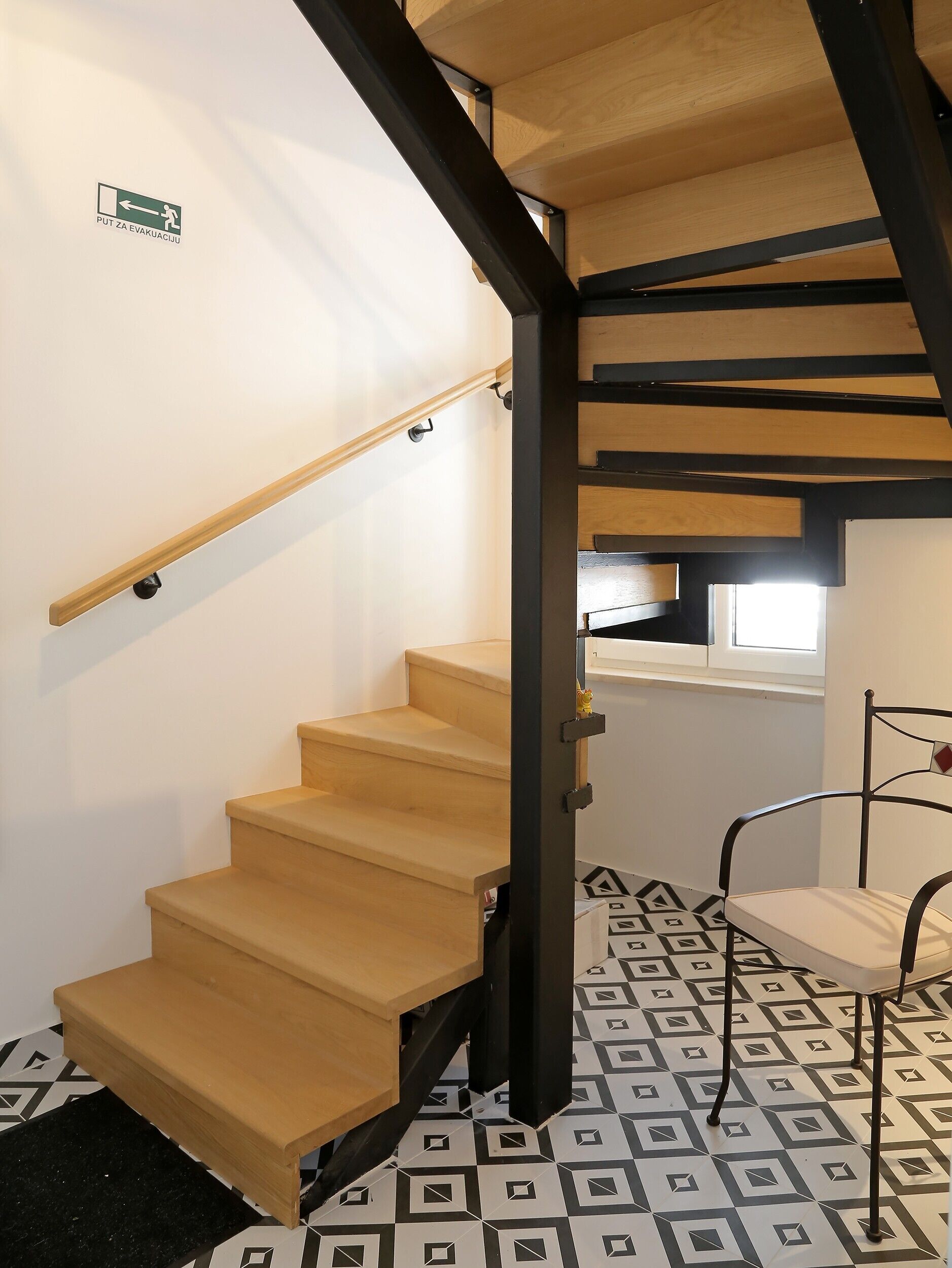 A wooden staircase with a black metal handrail, a chair, and patterned floor tiles.