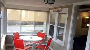 A covered patio area with a round table and four red chairs, large windows, and a view of the outdoors. - NUBBLE LIGHT VIEWS (York)