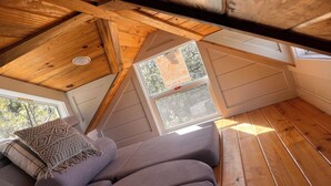 Ett sovrum i loftstil med ett sluttande tak, synliga träbjälkar, ett fönster med utsikt över träd och en grå soffa med ett vävt överkast i makramé.