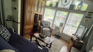Ein gemütliches Wohnzimmer mit einer Couch, einem Sessel, einem Tisch mit Obstschale und einem Fenster mit Jalousien.