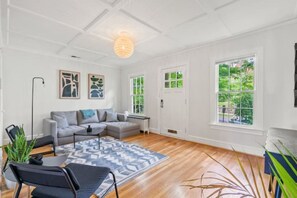 A modern living room with a sectional sofa, a coffee table, and a patterned rug. There are two windows with views of greenery outside.