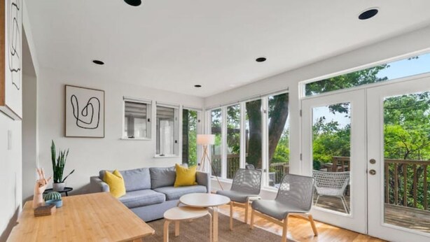 Ein großzügiger Wohnraum mit Holztisch, grauem Sofa und grauem Sessel. Ein großes Fenster bietet Blick ins Grüne.