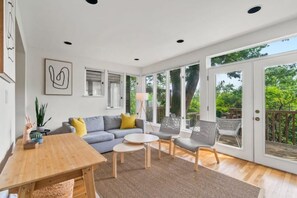A spacious living room with a wooden table, a gray sofa, and a gray armchair. There is a large window with a view of greenery outside. - Terraced Townhouse Getaway in Georgetown (Washington)
