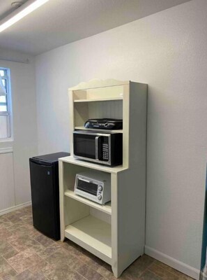 Compact kitchenette with shelving unit, microwave, and toaster oven, and a refrigerator.