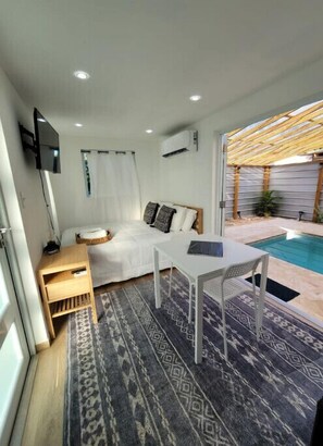A compact room with a bed, a small table, a TV, and a pool area with a pergola. - El Rinconcito Container Home Exp! Private Pool (Rincon)