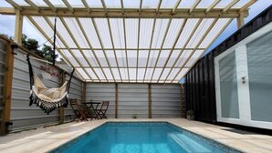 Una piscina exterior cubierta con una pérgola, una hamaca y una mesa con sillas.