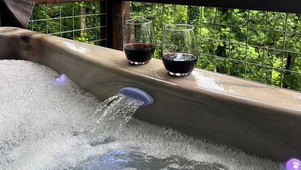 Un jacuzzi con dos copas de vino tinto en una terraza de madera, rodeado por una valla metálica y abundante vegetación.