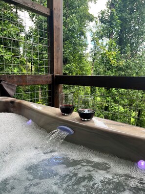 Een hottub met twee glazen rode wijn op een houten terras, omgeven door een metalen hek en weelderige begroeiing.