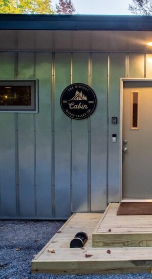 A metal building with a door and a window, a circular sign with the text "The Rideout Cabin 2021 Bears Valley, TX", and a wooden platform with a bench in front. - Scandinavian HideOut w/hot tub, mtn view+Starlink (Sevierville)