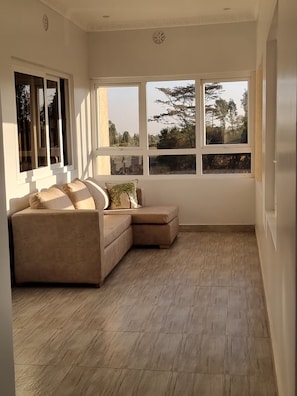 A corner room with a beige sectional sofa, large windows with a view of trees and buildings, and a wooden floor. - Casa Nadreya (Nakuru)