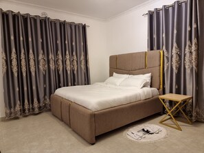 A bedroom with a large bed, curtains, a bedside table, and a small rug. - Casa Nadreya (Nakuru)