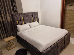 A bed with a tufted headboard, a wooden side table, and a small round table. - Casa Nadreya (Nakuru)