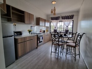 A modern kitchen with wooden cabinets, a stainless steel refrigerator, a microwave, and a dining area with metal chairs and a table. - Casa Nadreya (Nakuru)
