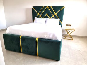 Una cama con cabecera y piecera tapizados en verde, ropa de cama blanca y una mesita de noche de madera con una bandeja de motivos dorados.