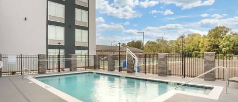 A swimming pool with a modern building in the background, a clear sky, and a fenced area.