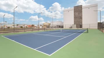 Una cancha de tenis al aire libre con red, rodeada por una valla, y contigua a un edificio con un ventanal grande y un muro de ladrillo.