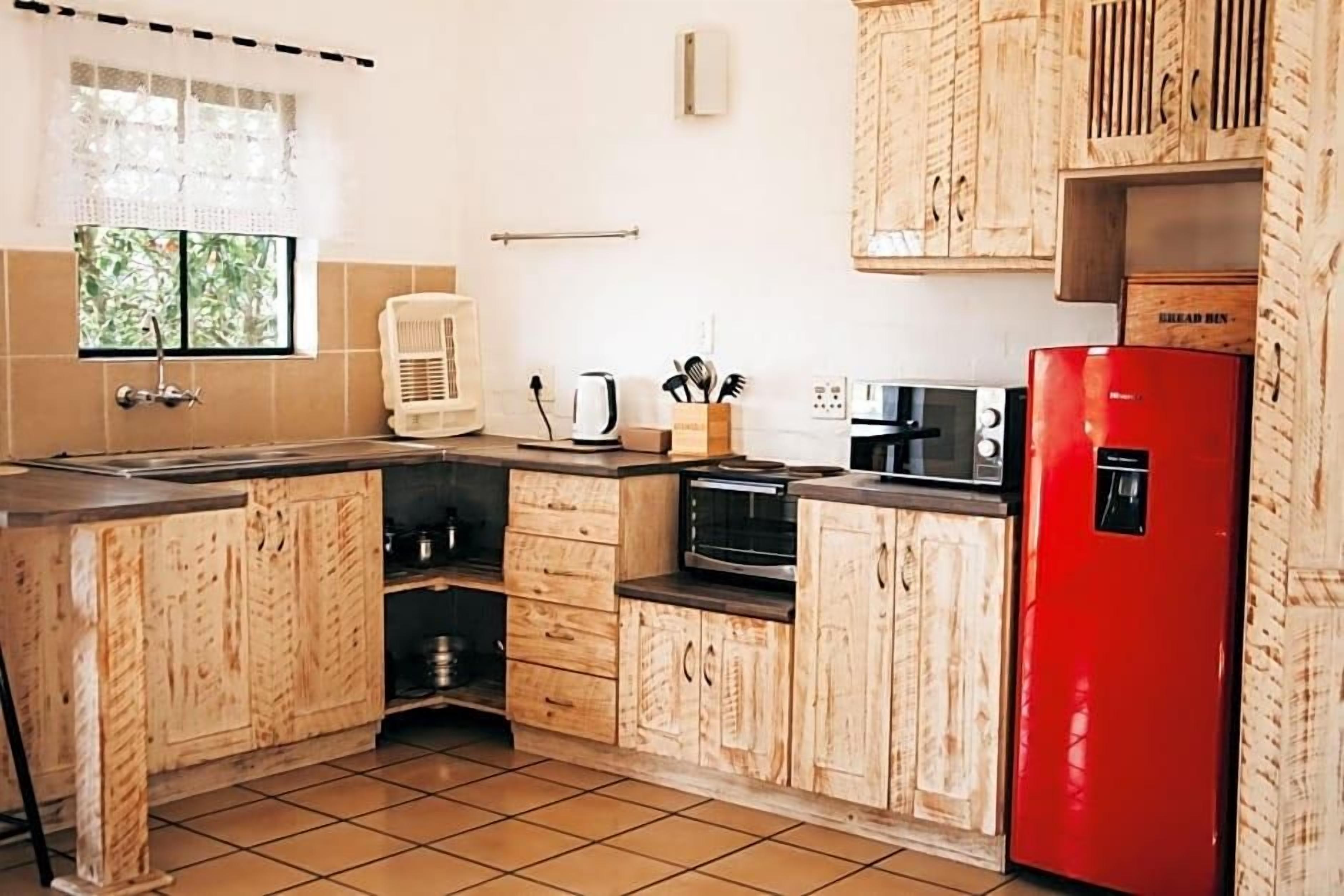 A kitchen with wooden cabinets, a red refrigerator, a kettle, and a window with a view of greenery.