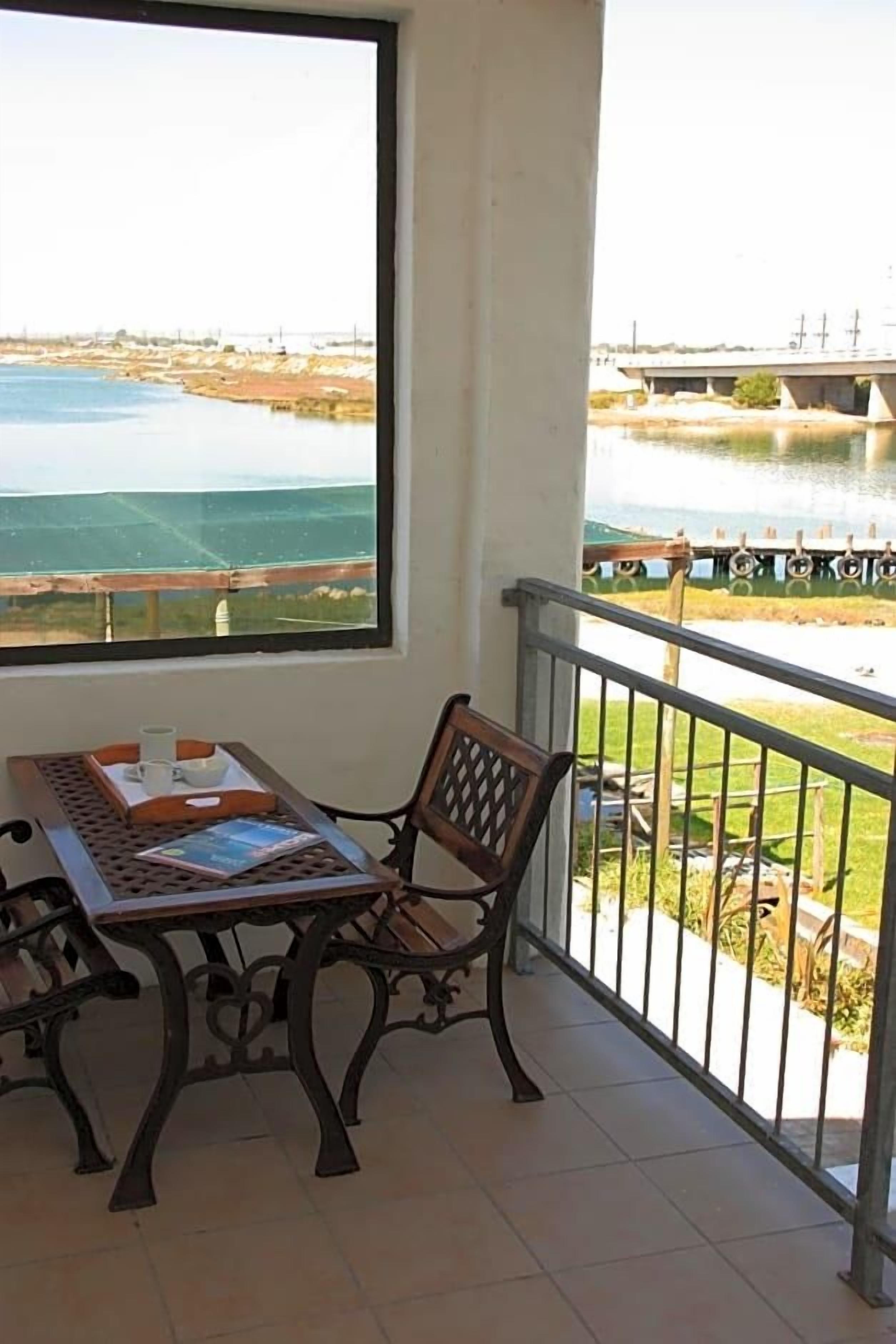 A balcony with a table and chair, a view of a body of water, and a bridge in the distance.