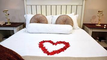 Una cama impecablemente hecha con un arreglo de pétalos de rosa roja en forma de corazón sobre el edredón, flanqueada por dos mesitas de noche con lámparas.