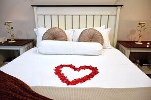 Una cama impecablemente hecha con un arreglo de pétalos de rosa roja en forma de corazón sobre el edredón, flanqueada por dos mesitas de noche con lámparas.
