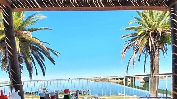 A table with food, drinks, and a hat, set under a thatched roof with palm trees and a view of the sea. - Riviera Hotel (Bergrivier)