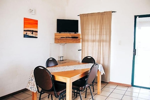 A dining area with a wooden table, four chairs, a television on a wooden stand, a painting on the wall, and a sliding glass door.