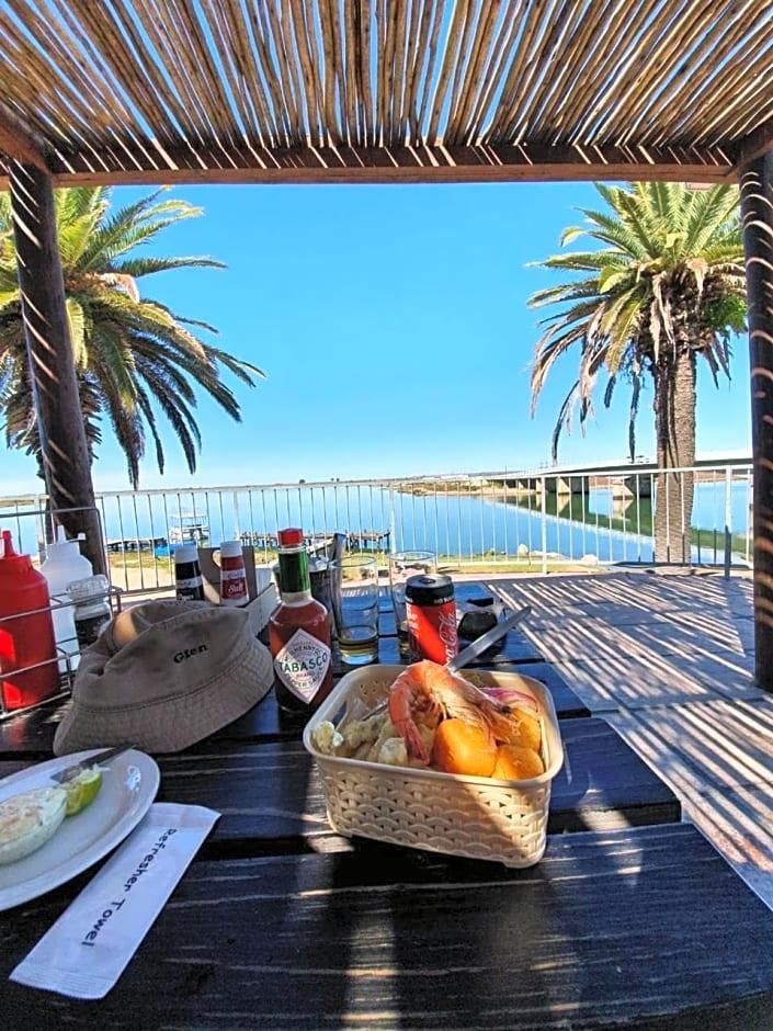 Une table dressée avec de la nourriture, des boissons et un chapeau, placée sous un toit de chaume, entourée de palmiers et offrant une vue sur la mer.