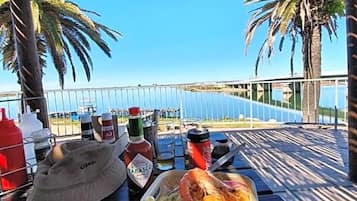 Una mesa con comida, bebidas y un sombrero, bajo un techo de paja con palmeras y vista al mar.