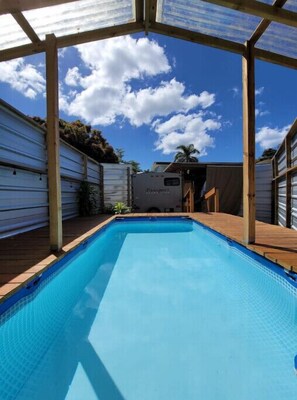 A swimming pool with a wooden deck, surrounded by covered structures and a clear blue sky. - El Rinconcito RV Experience 2.0! Private Pool (Rincón)